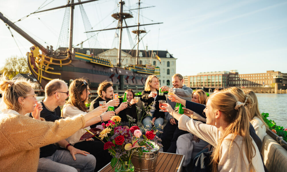 Group of people enjoying drinks on a canal boat in Amsterdam. A historic ship in the background.