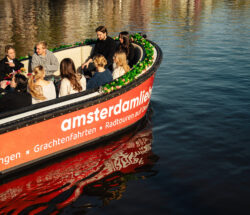A group of tourists on a boat tour in Amsterdam. The boat has an orange and white color scheme with green accents.