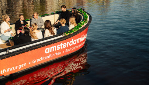 A group of tourists on a boat tour in Amsterdam. The boat has an orange and white color scheme with green accents.