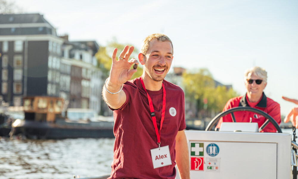 A smiling man waving on a boat in Amsterdam. A woman is sitting behind him.