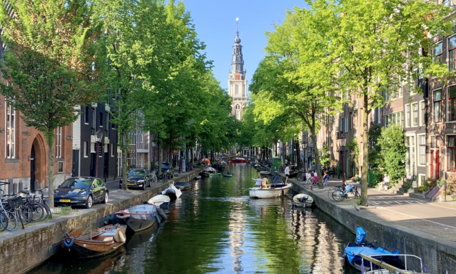 A serene canal with boats and lush green trees lining the waterway. The calm water reflects the vibrant colours of the scene.