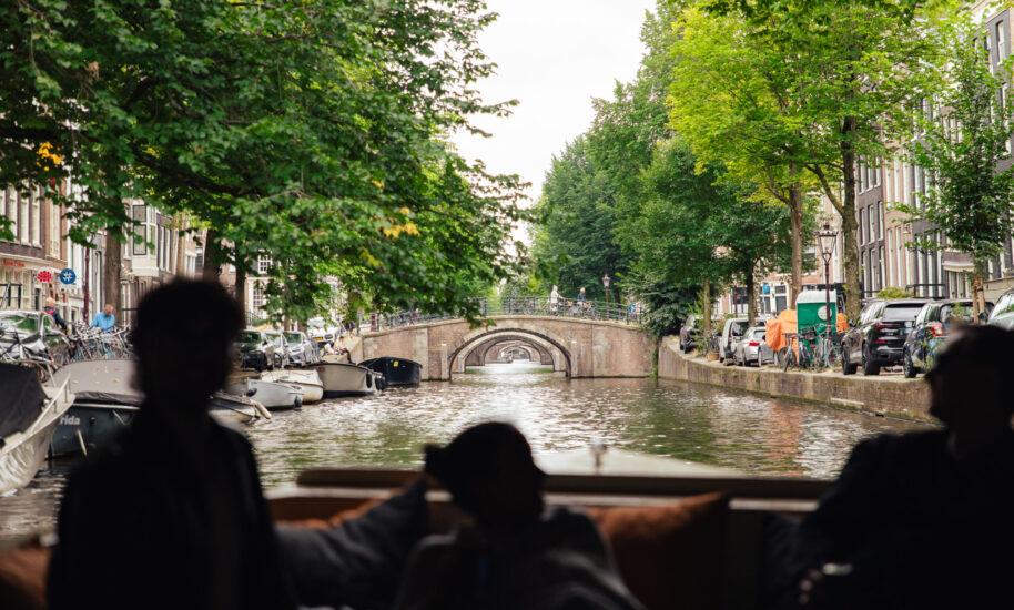 A scenic view of a canal in Amsterdam. The canal is lined with trees and features a bridge.