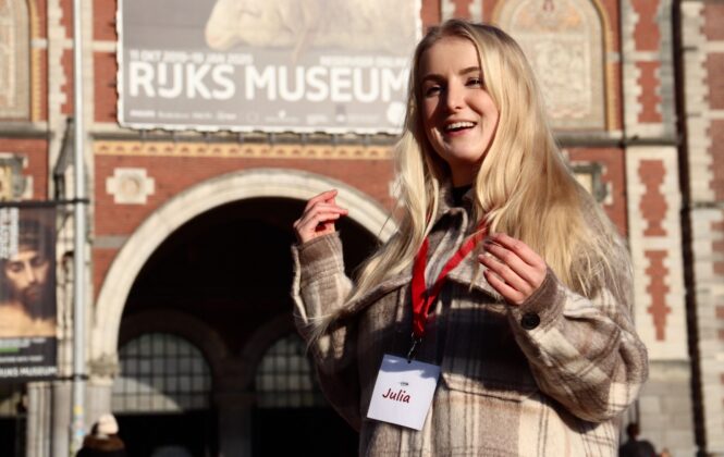 Blonde woman in beige jacket standing outside museum. She is wearing a red lanyard around her neck.