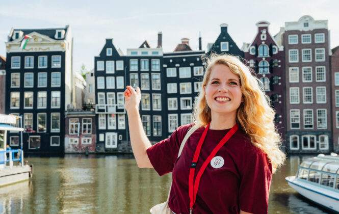 Tour guide Nina is smiling during a tour with Amsterdamliebe. She is standing in front of the famous dancing houses in Amsterdam city center and wearing a red shirt from Amsterdamliebe and a name tag around her neck.