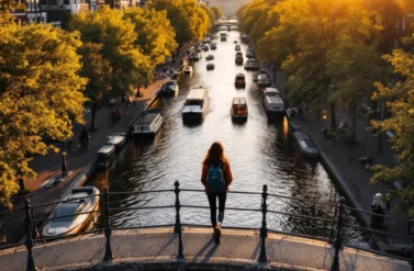 A woman stands on a bridge overlooking a busy canal. Boats and cars fill the waterway in Amsterdam.