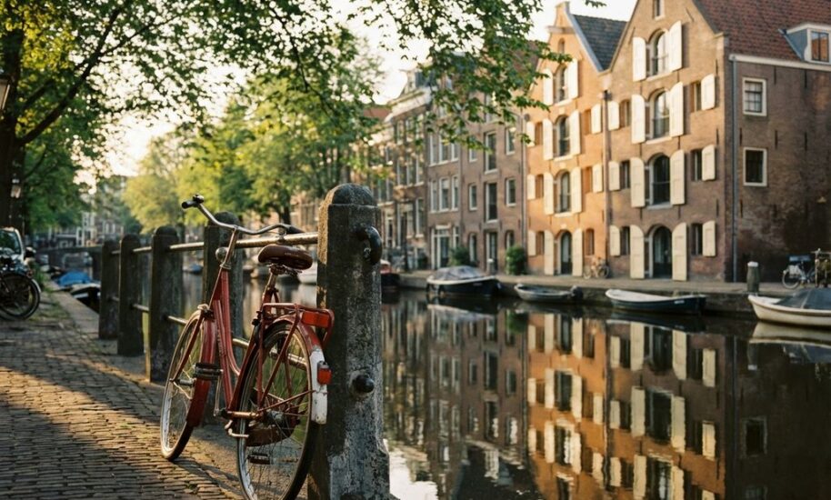 Red bicycle leaning against Amsterdam canal with traditional Dutch gabled houses reflected in still water at golden hour.