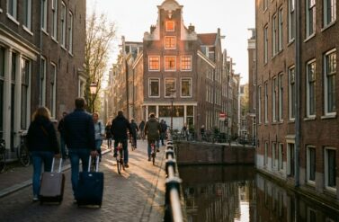 Amsterdam canal house with narrow gabled architecture and glowing windows, tourists with suitcases and cyclists on cobblestone street.