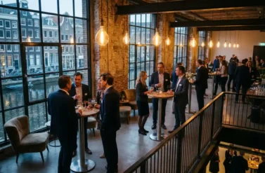 Office colleagues networking with cocktails at modern Amsterdam canal-side venue with industrial windows and warm lighting.