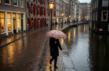 Person with colorful umbrella walking on wet cobblestone street in Amsterdam canal district during light rain at dusk