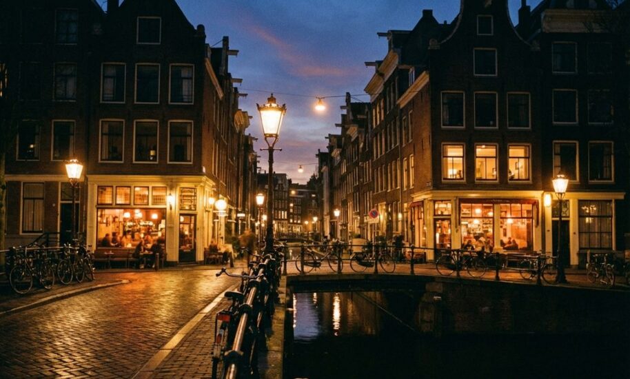 Amsterdam canal at twilight with historic gabled buildings, glowing café windows, street lamps reflecting in water, and bicycles