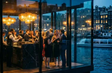 Elegant evening party at upscale Amsterdam canal-side venue with guests mingling, bartender serving drinks, city lights reflected in windows.