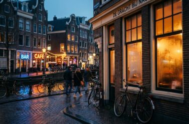 Amsterdam street corner at golden hour with warm café lights, wet cobblestones, bicycles, and historic brick buildings.
