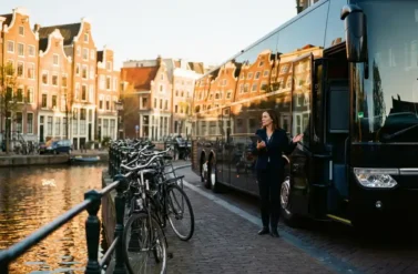 Black luxury coach bus parked beside Amsterdam canal with Dutch townhouses, event coordinator with tablet, bicycles nearby