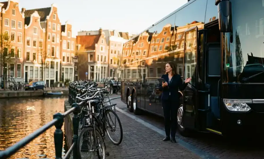 Black luxury coach bus parked beside Amsterdam canal with Dutch townhouses, event coordinator with tablet, bicycles nearby