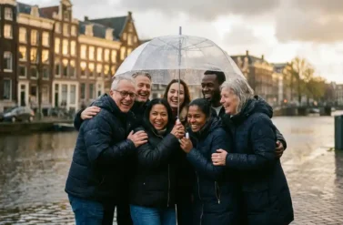 Diverse office colleagues sharing umbrella on rainy Amsterdam street with traditional Dutch canal houses in background