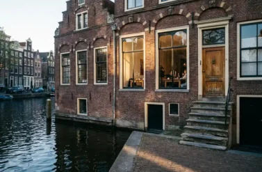 Weathered red brick Amsterdam canal house with ornate 17th-century gables reflected in dark water, warm light glowing from tall windows onto cobblestone quay.