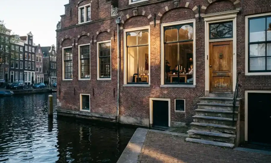 Weathered red brick Amsterdam canal house with ornate 17th-century gables reflected in dark water, warm light glowing from tall windows onto cobblestone quay.