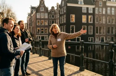 German tour guide leading tourists across Amsterdam canal bridge with historic gabled houses reflecting in water below