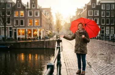 German tour guide with red umbrella on Amsterdam canal bridge during golden hour rain, historic gabled houses reflecting in wet cobblestones