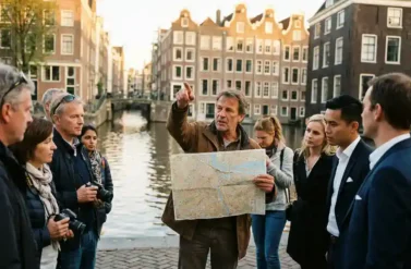 German tour guide with Amsterdam map explaining canal houses to attentive group of tourists and organizers beside historic Dutch grachten