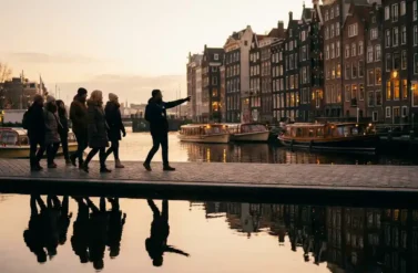 Tourists walking along Amsterdam canal during golden hour with historic gabled houses, tour guide, and peaceful boats reflected in calm water