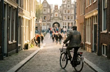 Narrow cobblestone alley in Amsterdam with cyclist riding toward distant crowds, traditional gabled canal houses lining both sides in golden hour light.