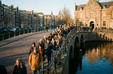 Long queue of tourists winding along Amsterdam's cobblestone street past canal-side gabled houses toward museum entrance in golden afternoon light
