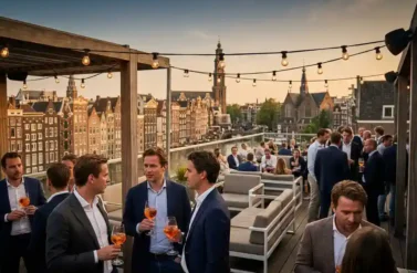 Professionals enjoying cocktails on Amsterdam rooftop terrace at golden hour with canal houses and church spires in background