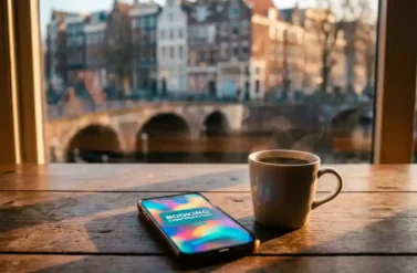 Smartphone with colorful booking confirmation screen on wooden café table next to steaming coffee cup, Amsterdam canal houses visible through window in background