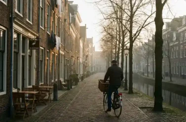 Cyclist with wicker basket rides cobblestone street past Dutch brick houses with canal and morning mist in golden light