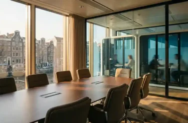 Modern conference room with polished wooden table, glass-walled breakout pods, and Amsterdam canal view through large windows with warm afternoon sunlight