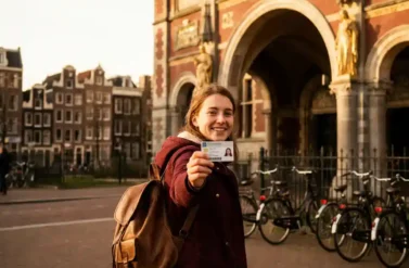 Young student with backpack holds student ID card while smiling at Rijksmuseum entrance, Amsterdam canal houses and bicycles in background
