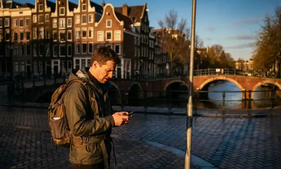 Tourist checking smartphone at empty Amsterdam canal with gabled houses and red brick bridge in golden hour light