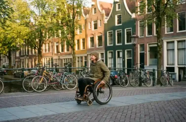 Person in wheelchair navigating wide accessible pathway along Amsterdam canal with historic gabled houses and parked bicycles