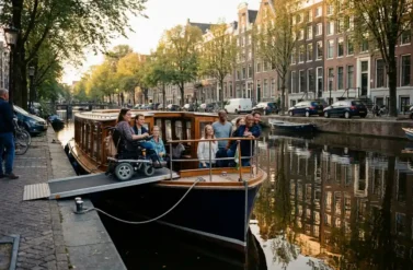 Wheelchair-accessible canal boat with ramp on Amsterdam's Herengracht canal, diverse tourists including wheelchair user enjoying historic gabled houses reflecting in water during golden afternoon light.